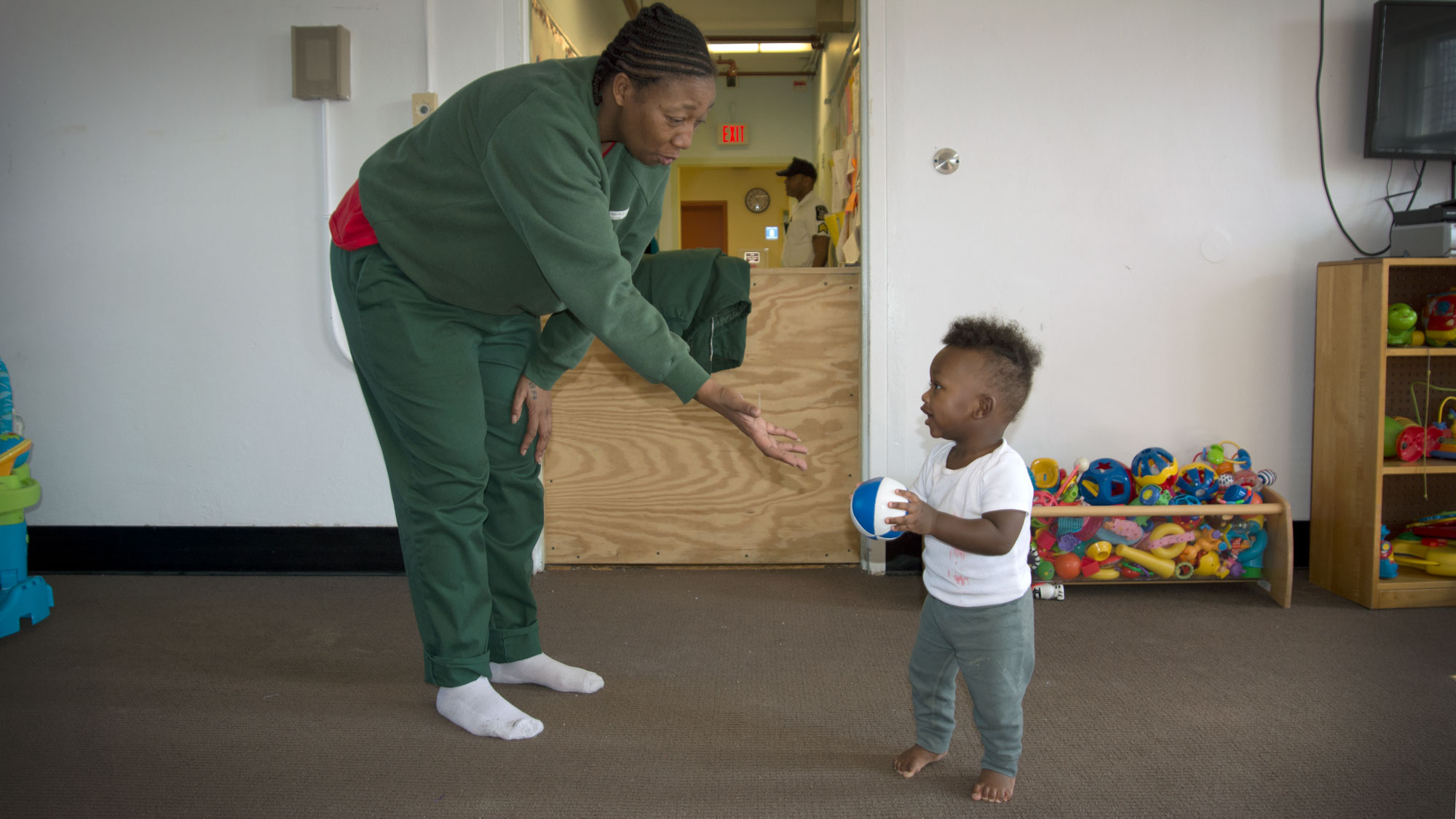 A woman holds out her hand to a toddler in a playroom. She is dressed in an olive green uniform.
